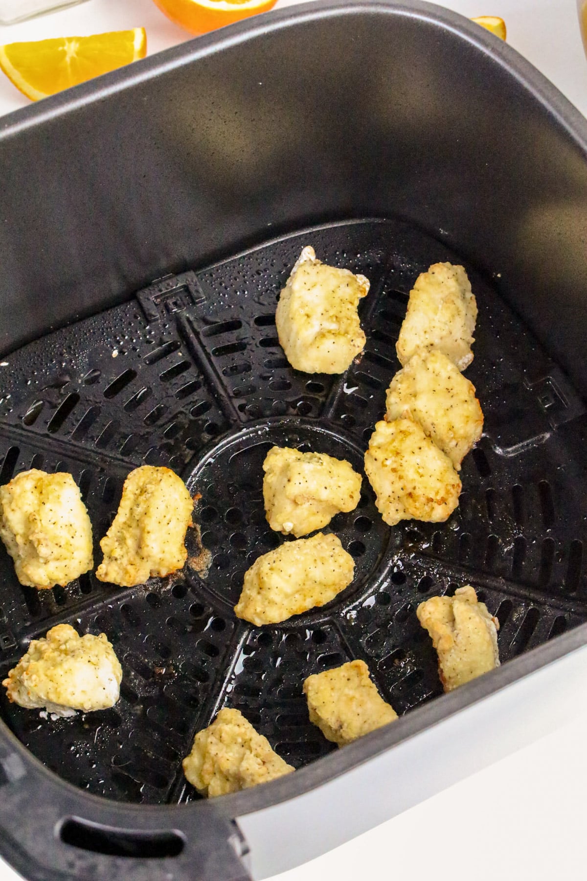 Chicken nuggets cooking in an air fryer basket, with more pieces on the side along with orange slices.