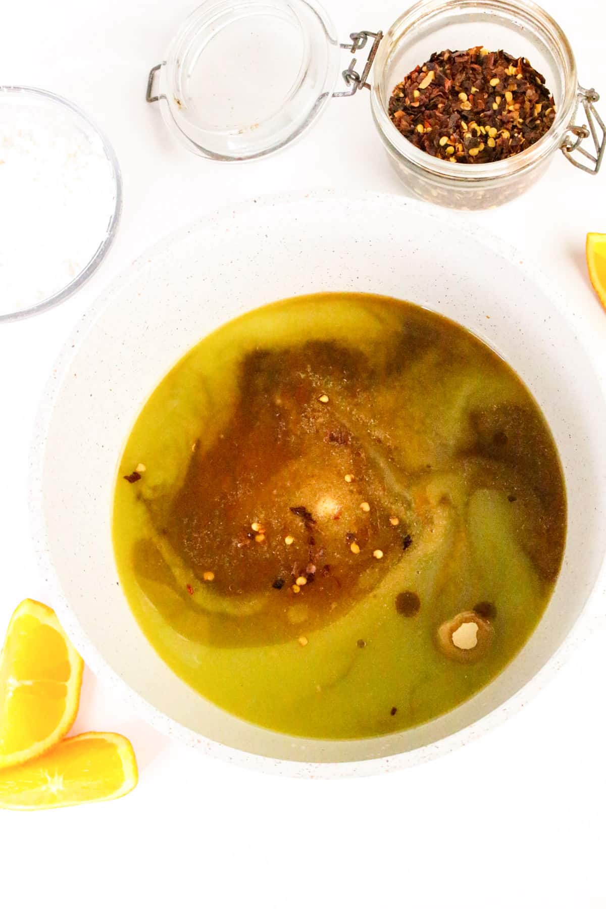Bowl of sauce with spices, surrounded by orange slices, chili flakes, and a jar of powder on a white background.