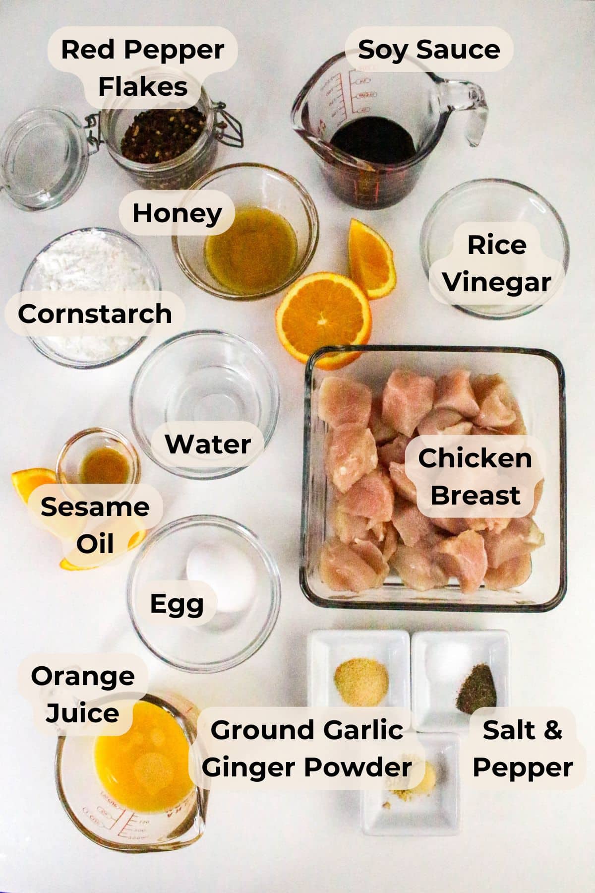 Labeled ingredients for Air Fryer Orange Chicken recipe arranged neatly on a white surface in bowls and containers.