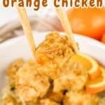 Chopsticks holding a piece of orange chicken over a bowl of rice, with orange slices in the background.
