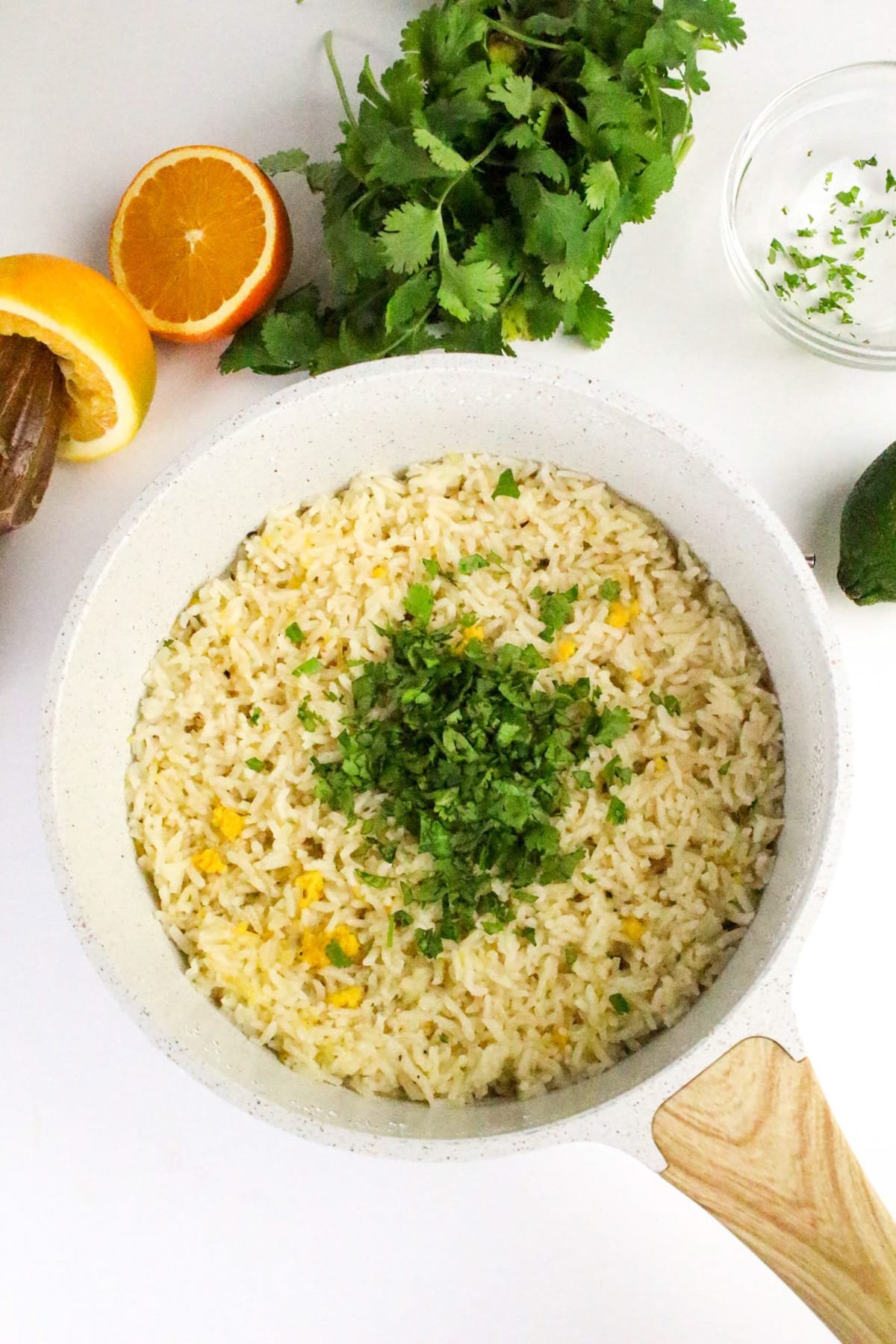 Rice dish garnished with cilantro in a pan, surrounded by a halved orange, cilantro bunch, avocado, and glass bowl.