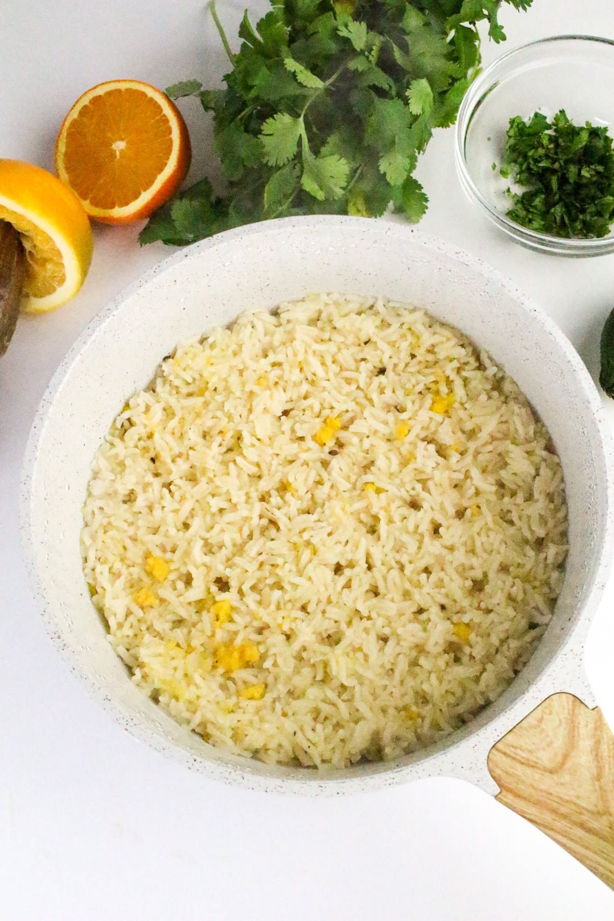 A pot of cooked rice with herbs, surrounded by cilantro, an orange half, and a small bowl of chopped herbs.