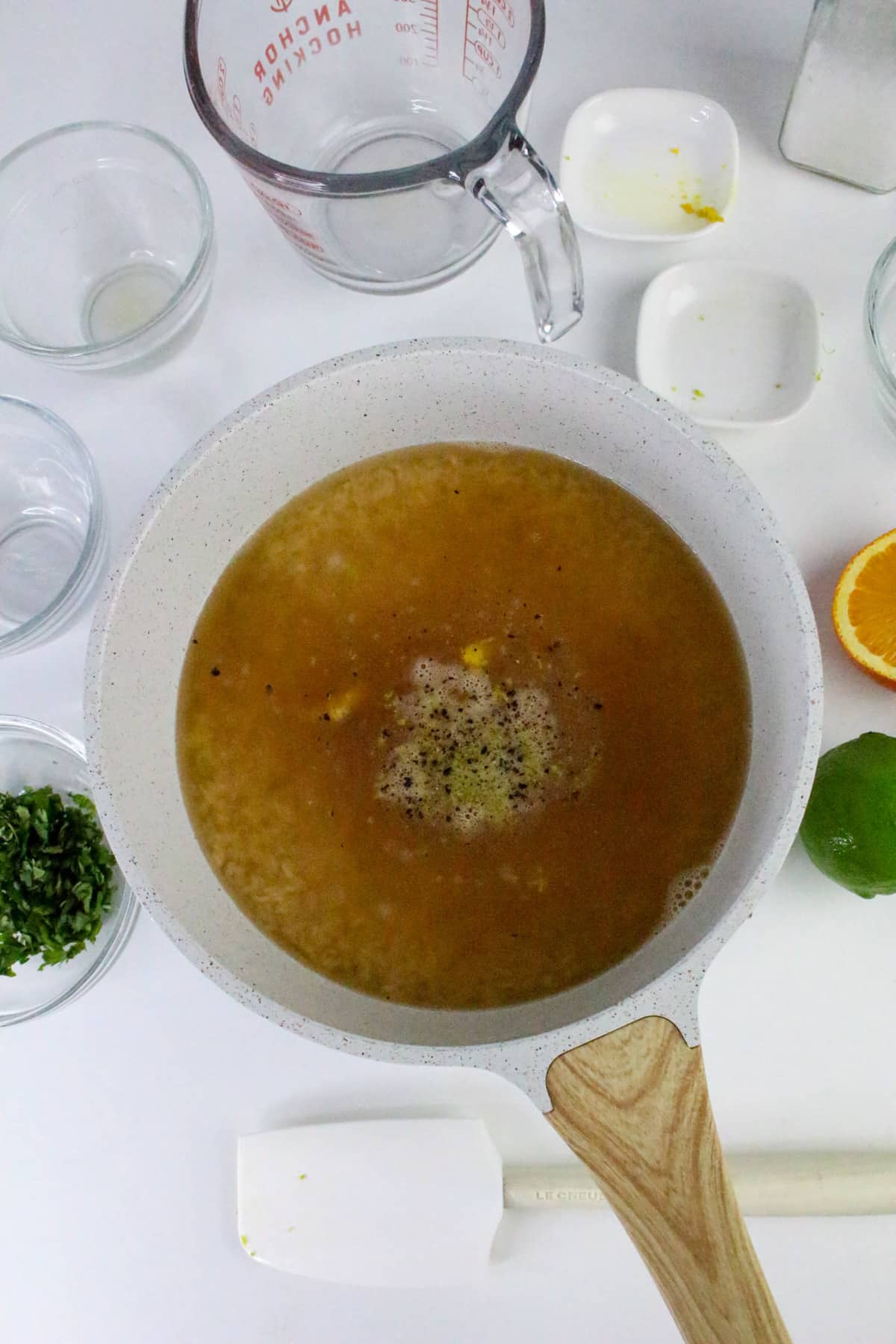 Sauce in a pan beside lemon, lime, orange, chopped herbs, spatula, and measuring cups on a white surface.