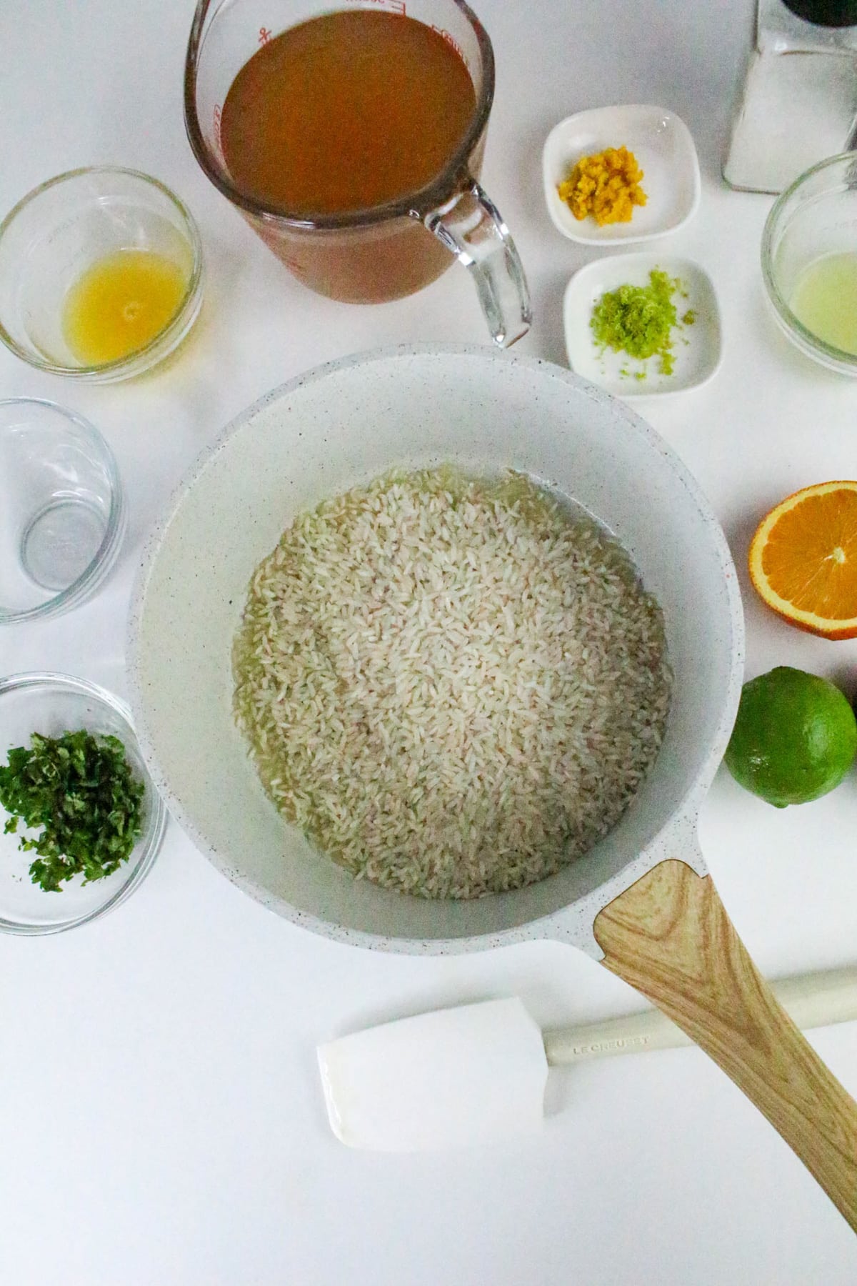 White pot with rice surrounded by bowls and plates of ingredients like broth, citrus zest, juice, and herbs.