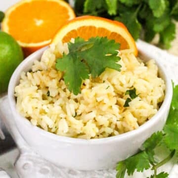 Bowl of rice garnished with cilantro, surrounded by orange slices, lime, and cilantro bunch.