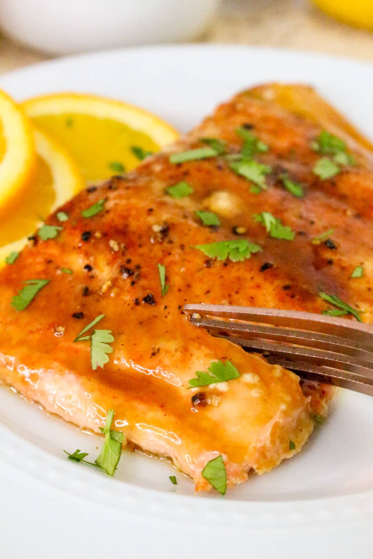 Close-up of glazed salmon on a white plate, garnished with herbs and lemon slices, with a fork cutting into it.