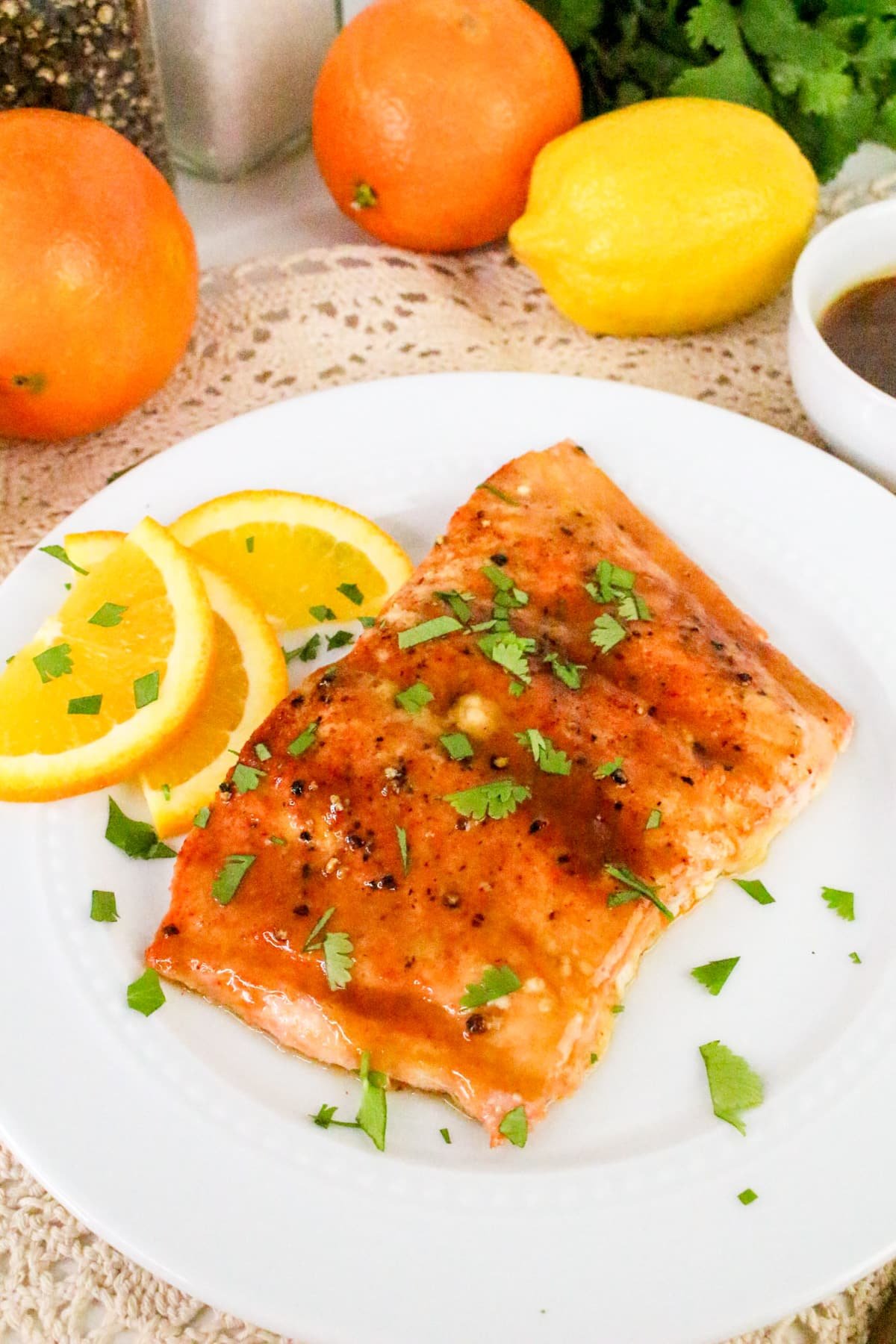 Grilled salmon topped with herbs, served with orange slices on a white plate, surrounded by citrus fruits.