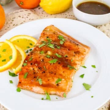 Grilled salmon garnished with herbs and served with orange slices on a white plate, with sauce in a small bowl nearby.