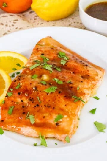 Grilled salmon garnished with herbs and served with orange slices on a white plate, with sauce in a small bowl nearby.