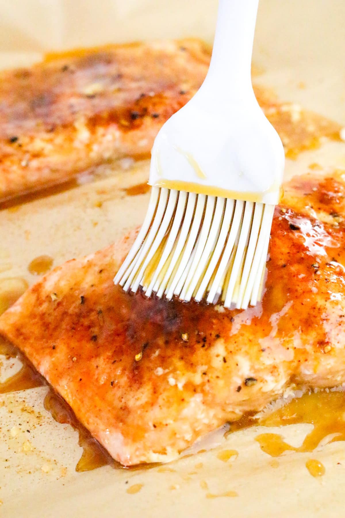 A white brush applies glaze to cooked salmon fillets on parchment paper.