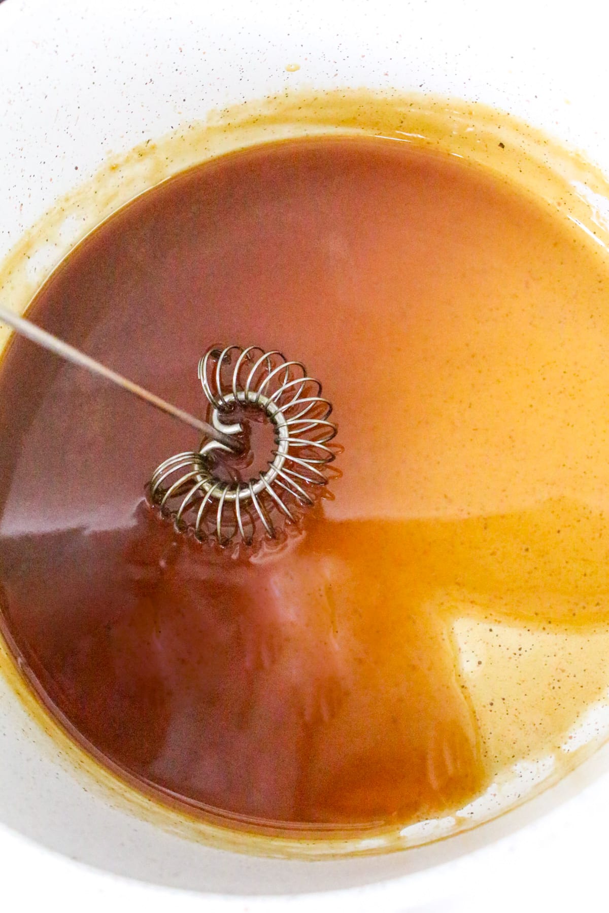 A whisk stirring caramel-colored liquid in a white bowl.