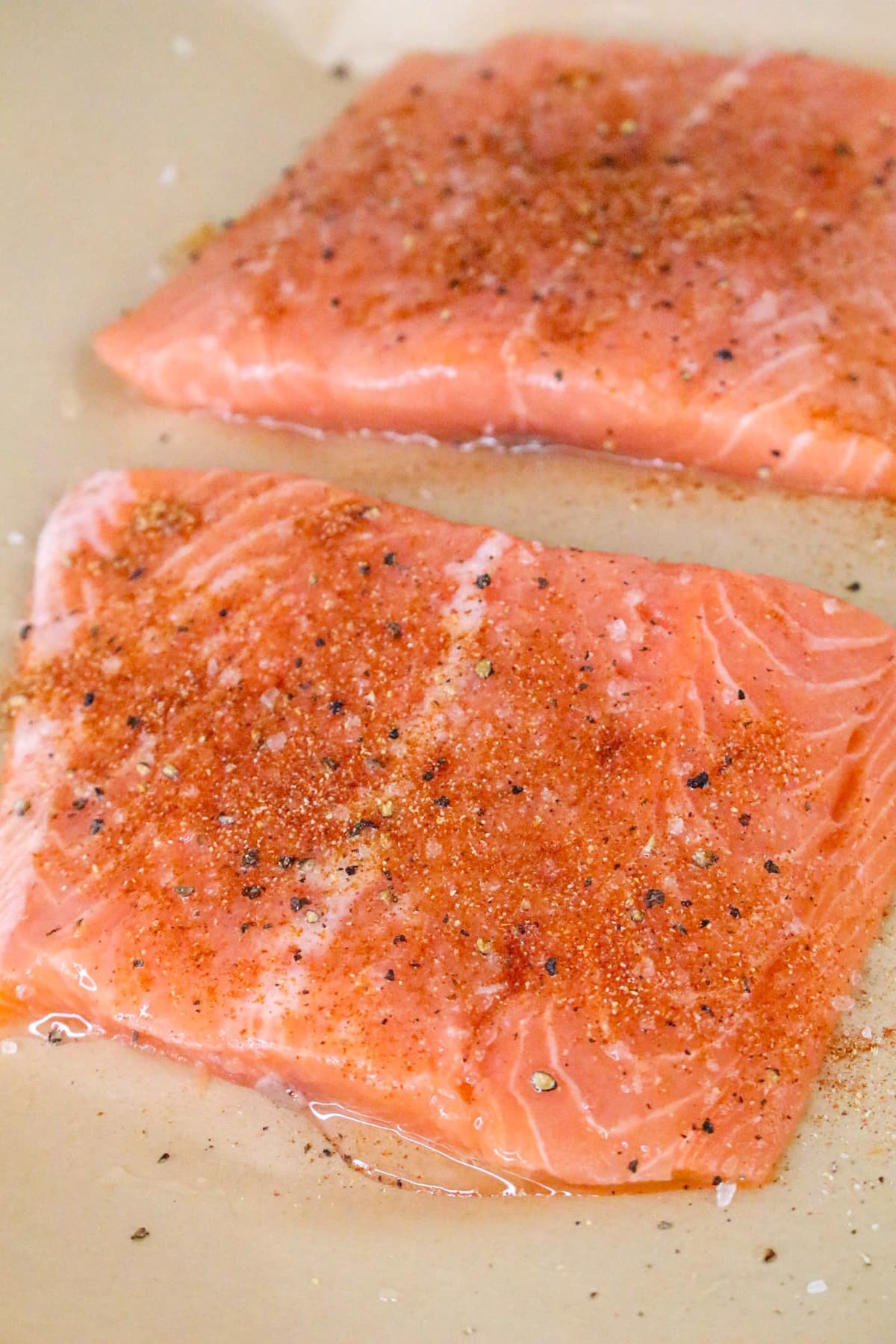 Two raw salmon fillets seasoned with pepper and spices on a beige surface.