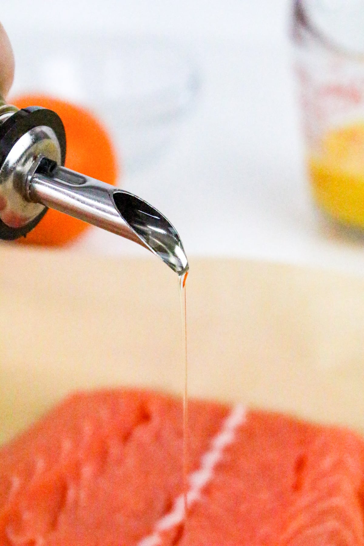 Close-up of oil being poured onto a raw salmon fillet, with a blurry background of an orange and a measuring cup.