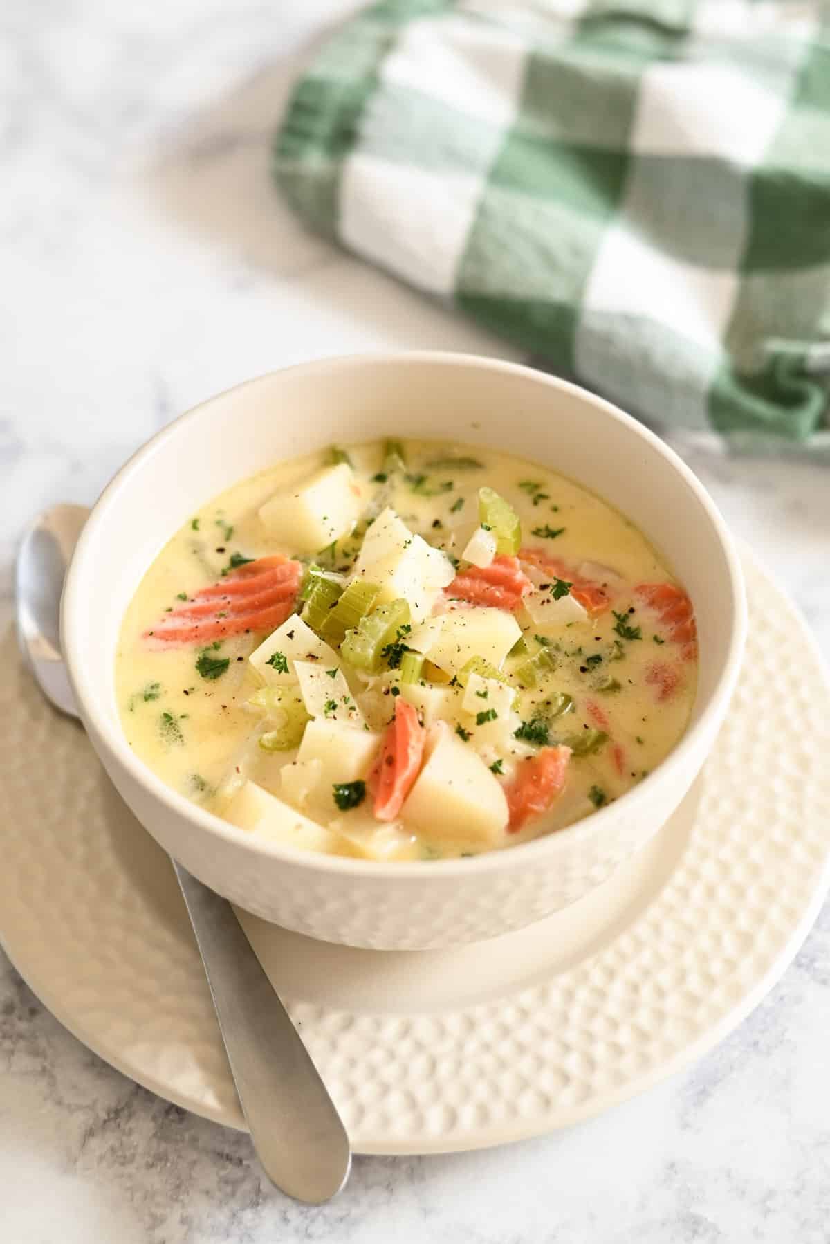 A bowl of creamy soup with vegetables, garnished with herbs, on a plate with a spoon, near a green checkered cloth.