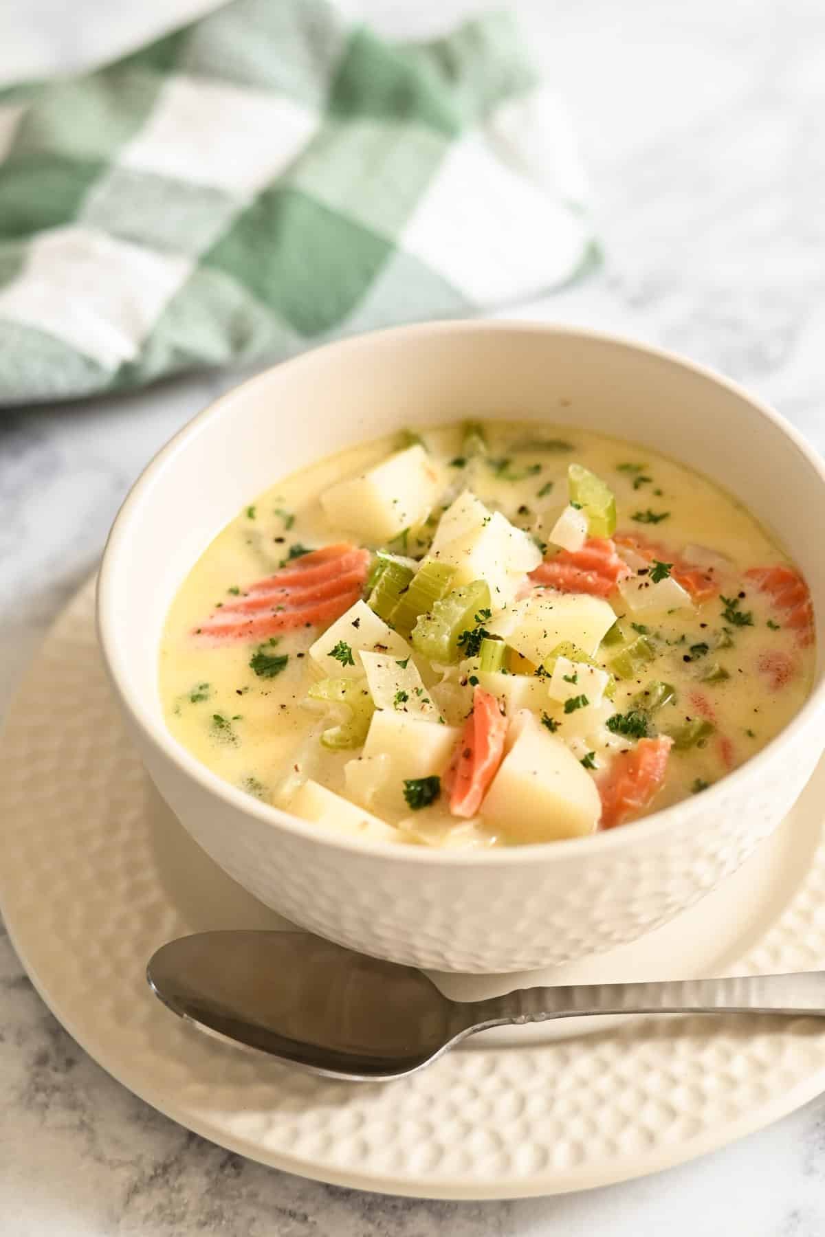 A bowl of creamy soup with vegetables and herbs, next to a green-checkered napkin and a spoon on the table.