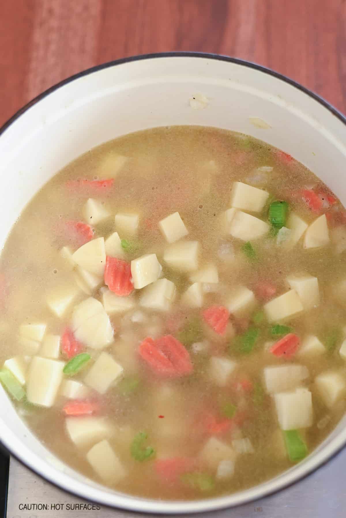 Pot of soup with diced potatoes, carrots, and celery in broth on a stovetop.