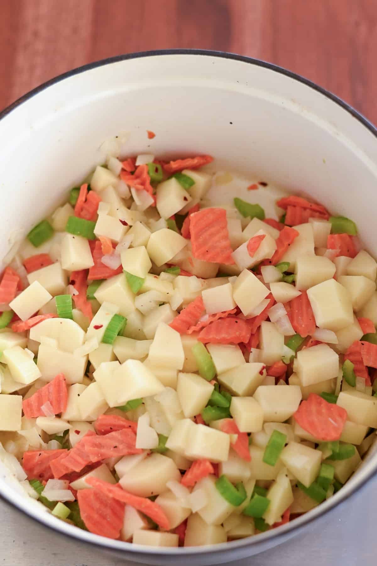 Chopped potatoes, carrots, and celery in a white pot on a wooden surface.