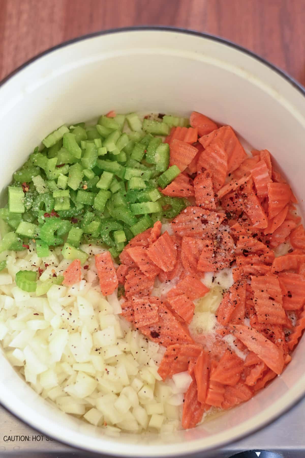 Pot with diced onions, celery, and carrots on a wooden surface.