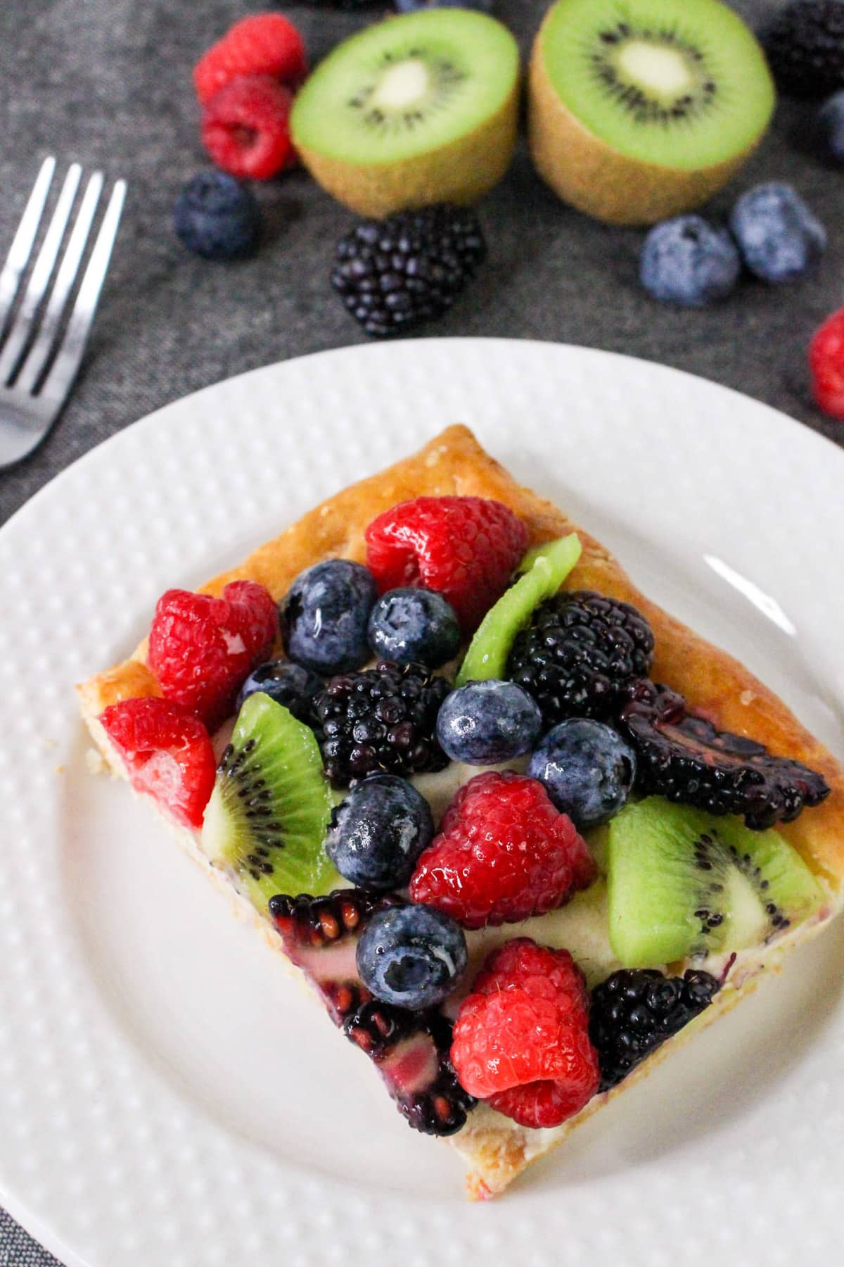 A fruit pastry slice topped with kiwi, raspberries, blueberries, and blackberries on a white plate.