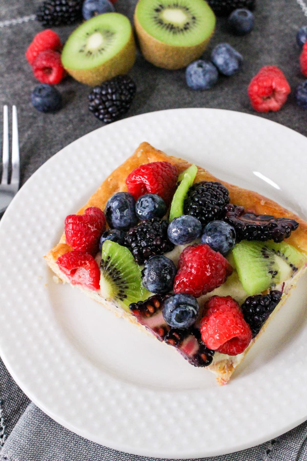 A slice of fruit tart with kiwi, raspberries, blackberries, and blueberries on a white plate.