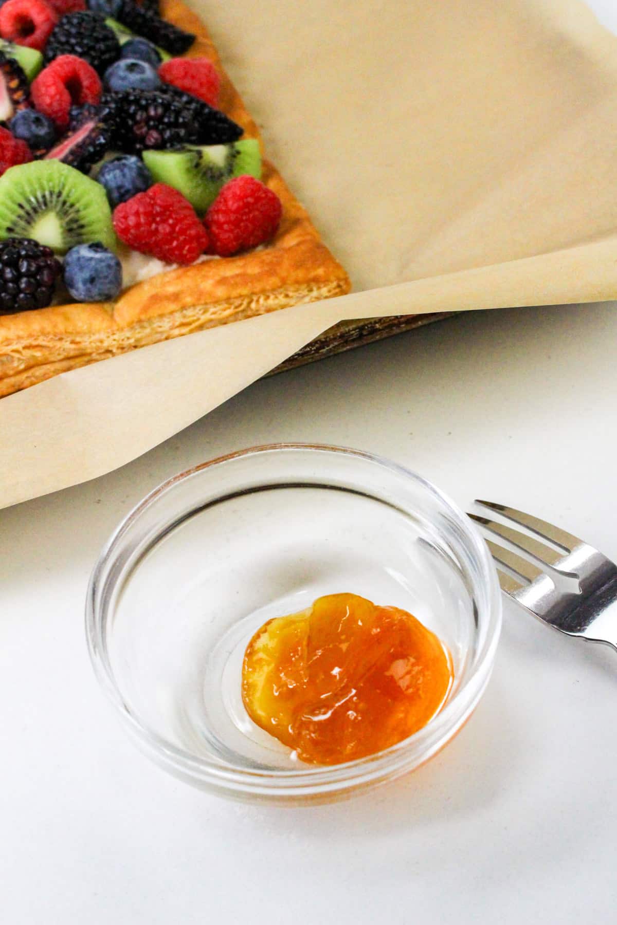 Fruit tart with kiwi, berries, and peaches on parchment paper; small bowl of jam and fork nearby.