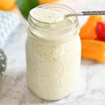 A jar of creamy ranch dressing with herbs, a spoon resting on top, on a marble surface with bell peppers in the background.