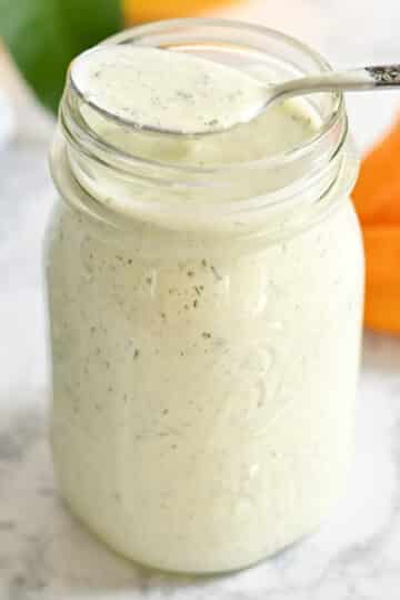 A jar of creamy ranch dressing with herbs, a spoon resting on top, on a marble surface with bell peppers in the background.