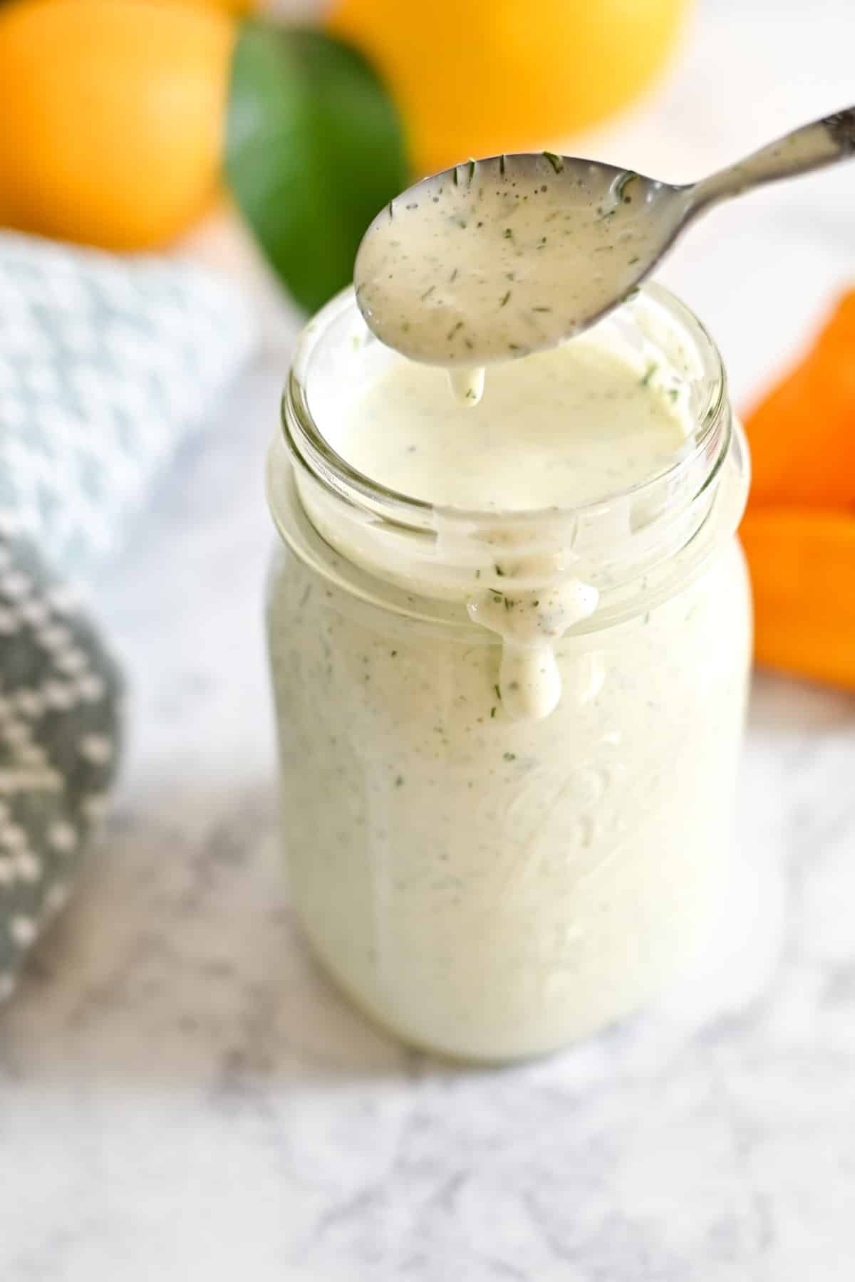 Spoon dripping white dressing into a jar on a marble surface, with oranges and cloth in the background.