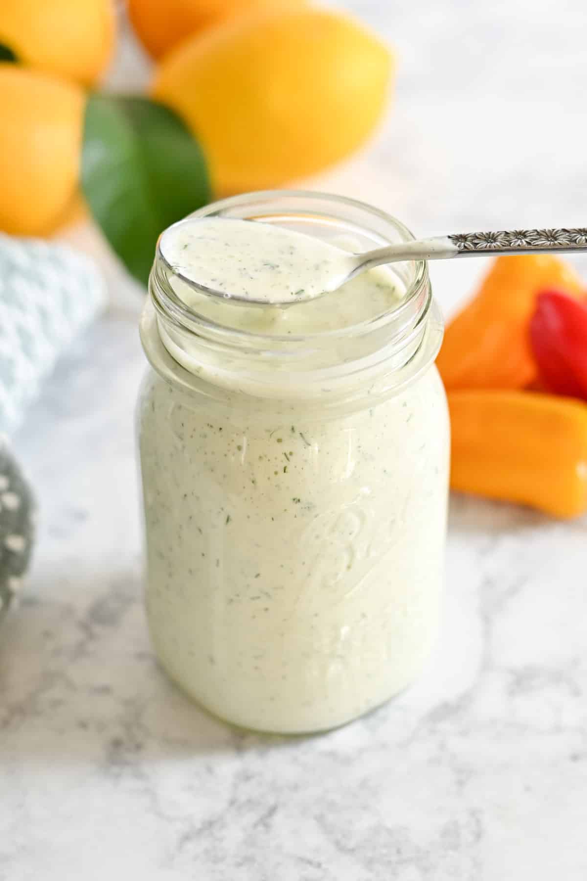 Jar filled with creamy ranch dressing, spoon inside, surrounded by colorful bell peppers and lemons on a marble surface.