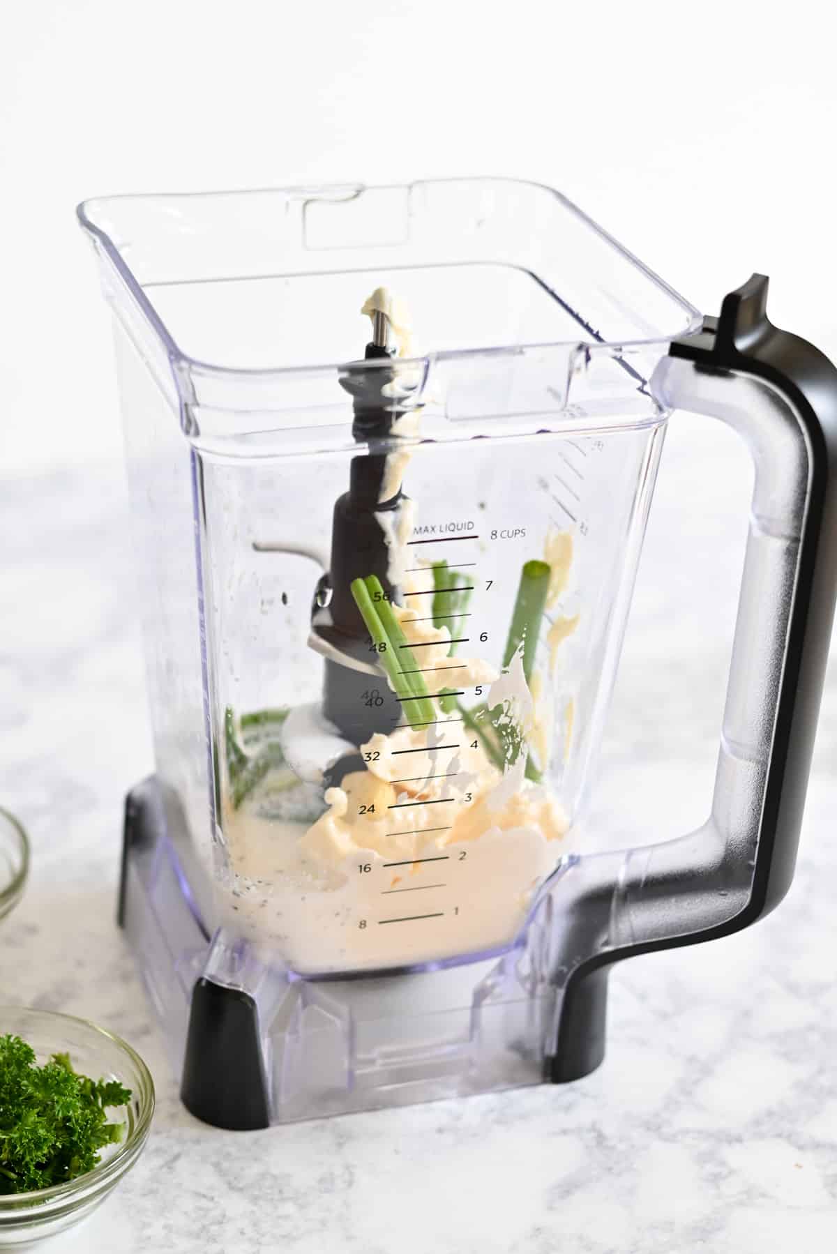 Blender with mayonnaise and green onions inside on a marble countertop, beside bowls of fresh herbs.