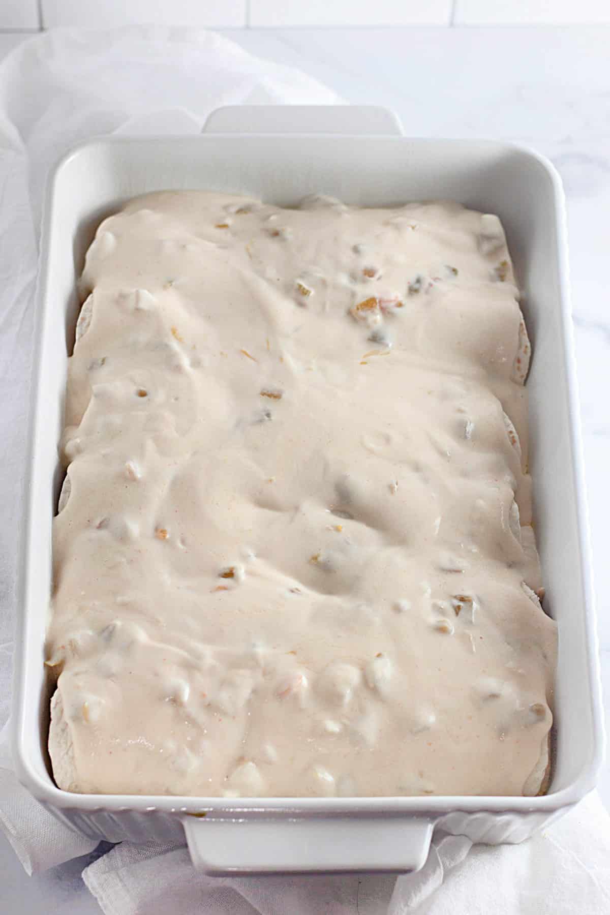 A white baking dish filled with uncooked, sauce-covered enchiladas.
