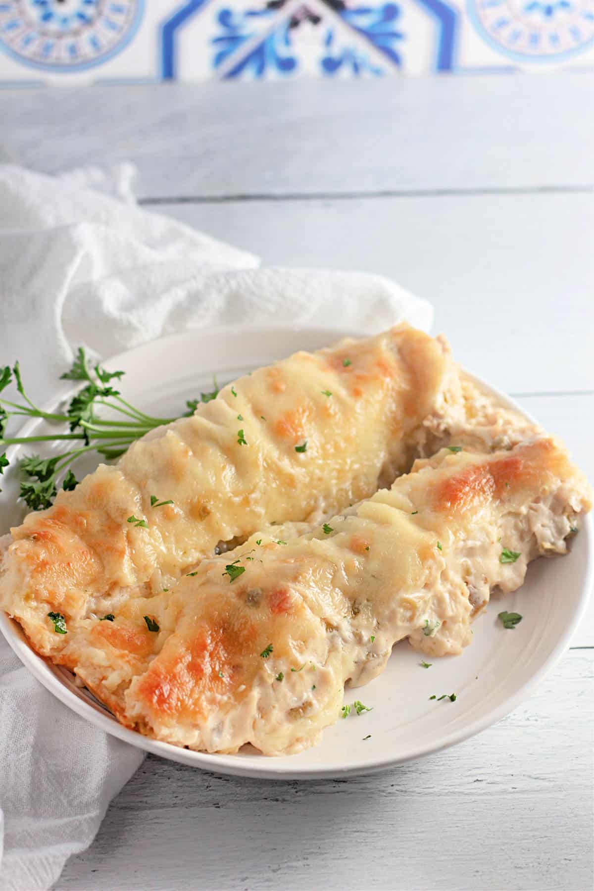 Two creamy chicken enchiladas topped with melted cheese on a white plate, garnished with chopped herbs.