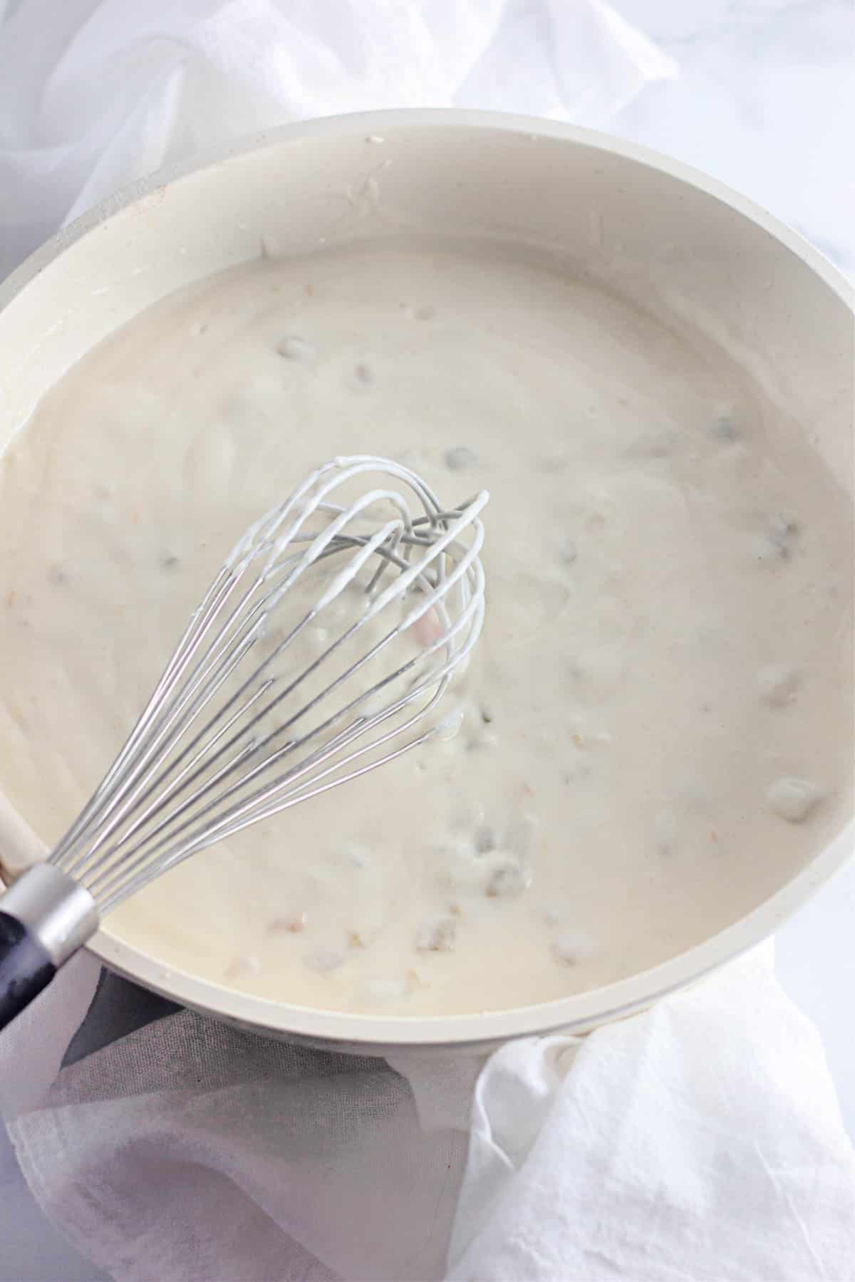 Whisk stirring creamy white sauce with small chunks in a white skillet on a white cloth.