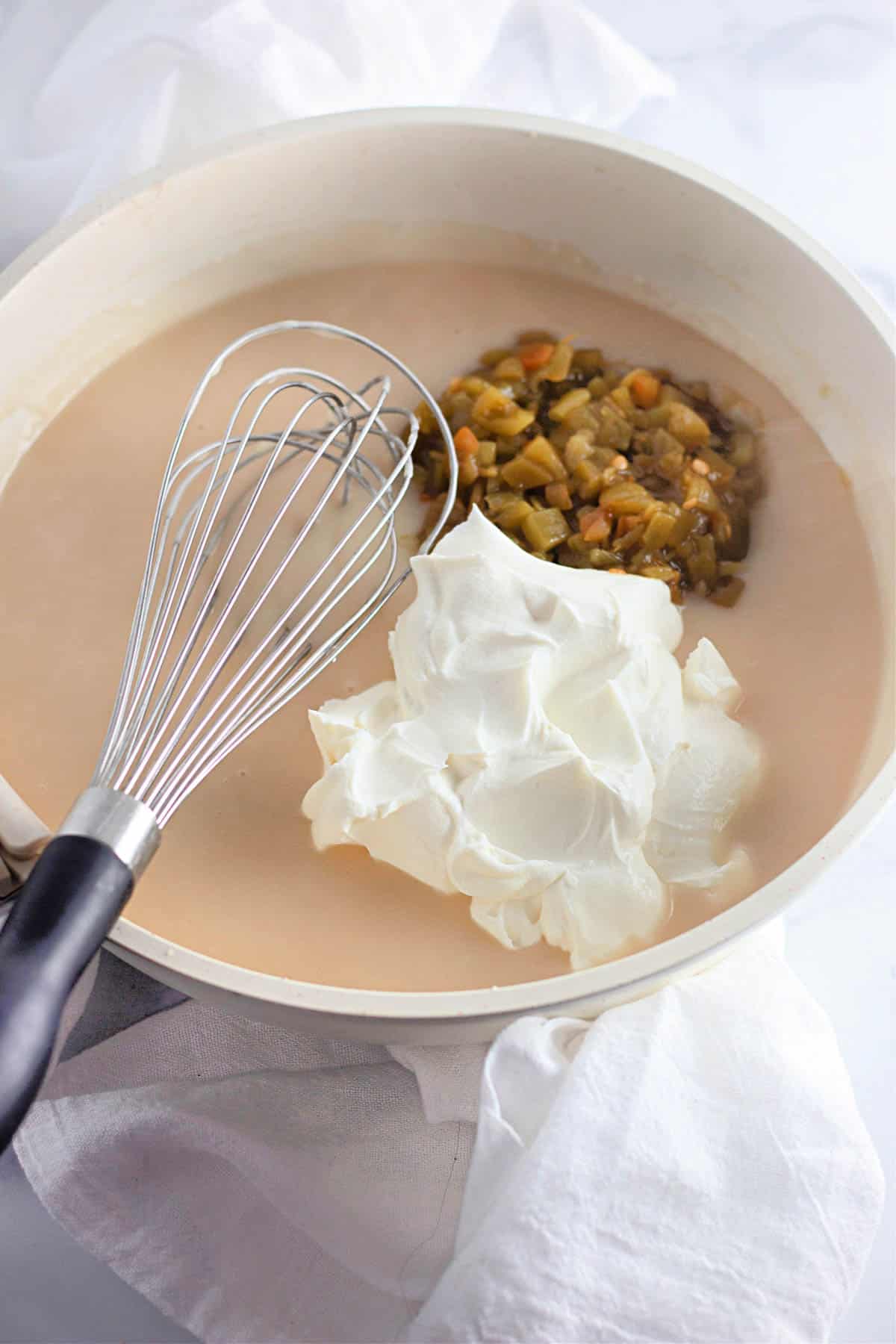Bowl with roux base, topped with chopped green chiles and a dollop of sour cream, with a whisk beside it.