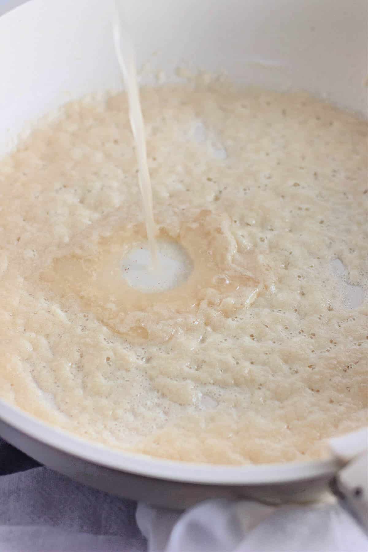 Liquid being poured into a white pan with bubbling beige sauce, creating a small splash in the center.