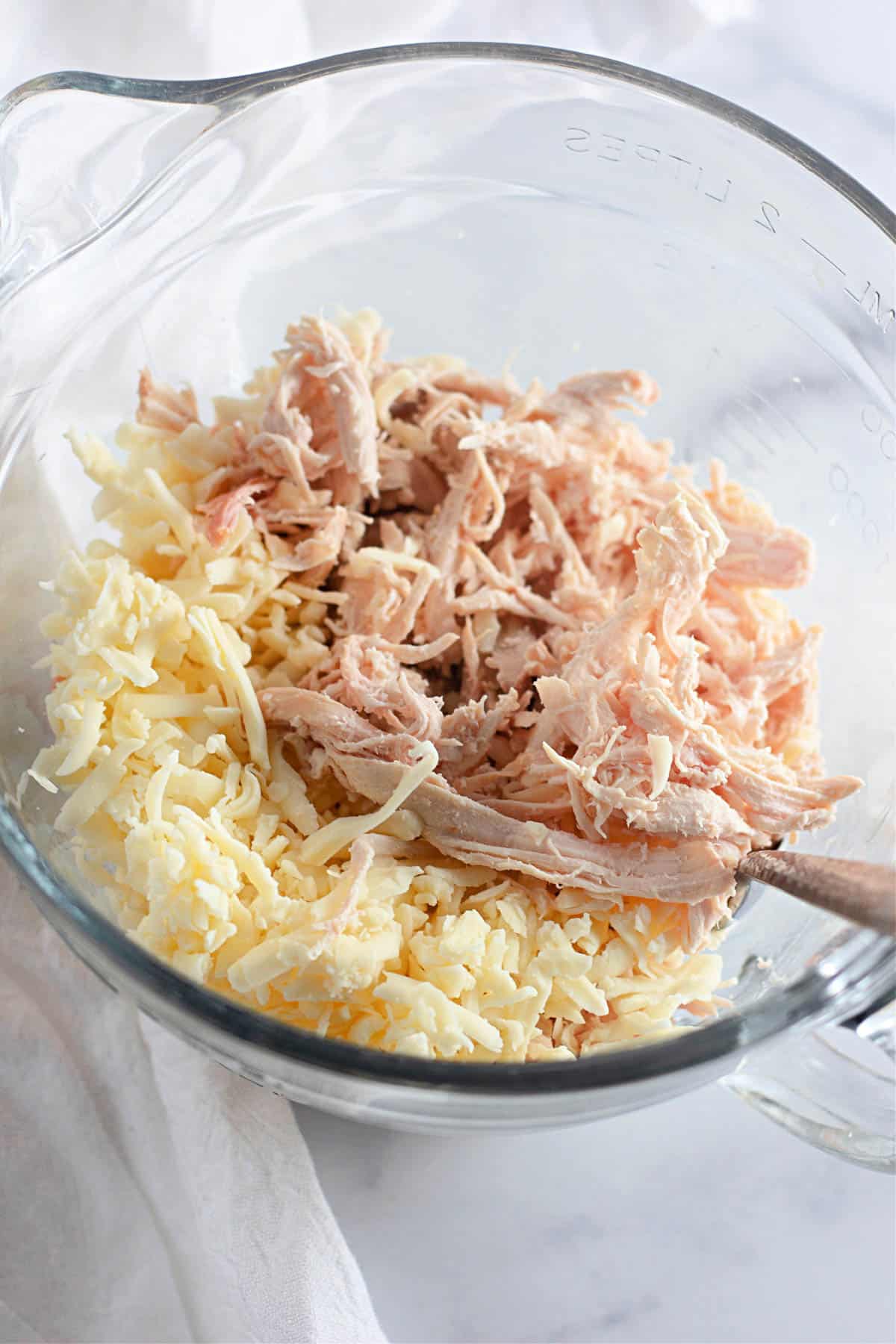 Shredded chicken and grated cheese in a clear glass mixing bowl on a white surface.