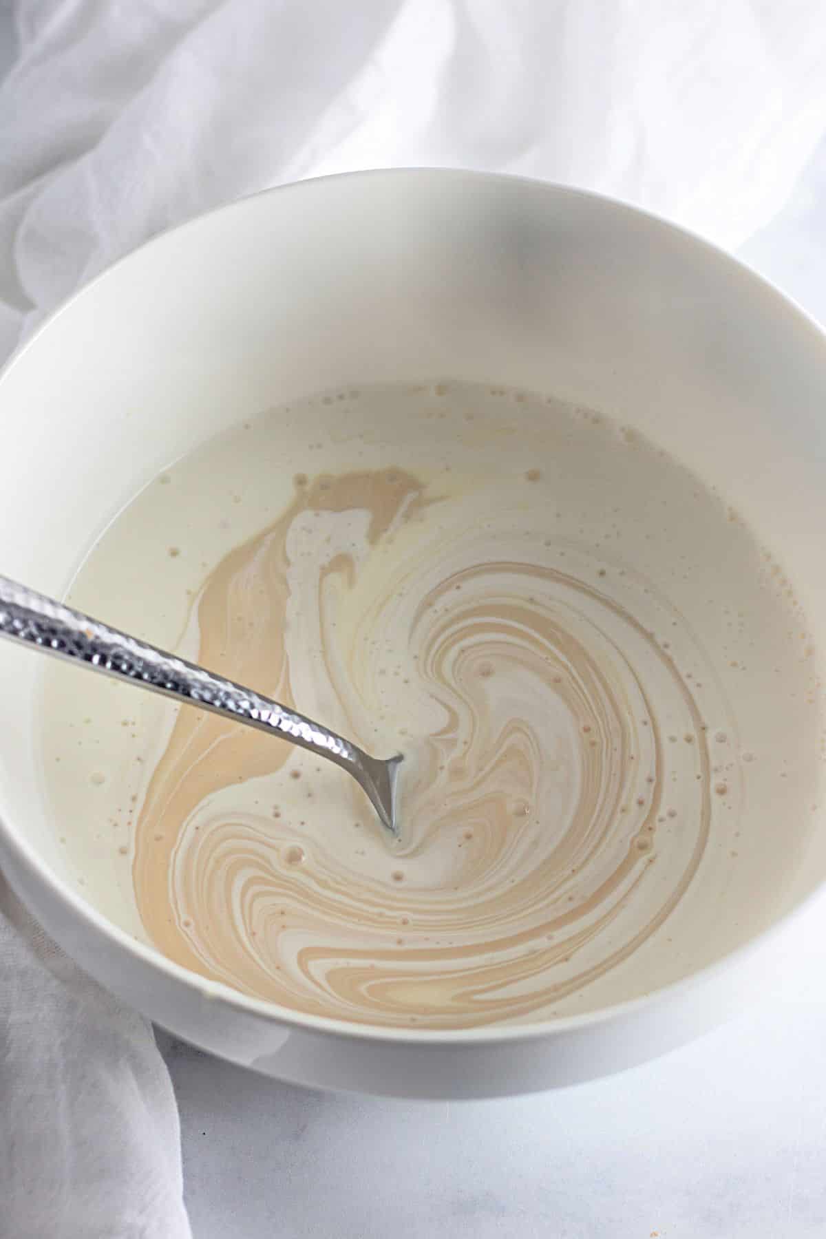 A spoon in a bowl of cream with swirled tan liquid, set on a white surface with a white cloth nearby.