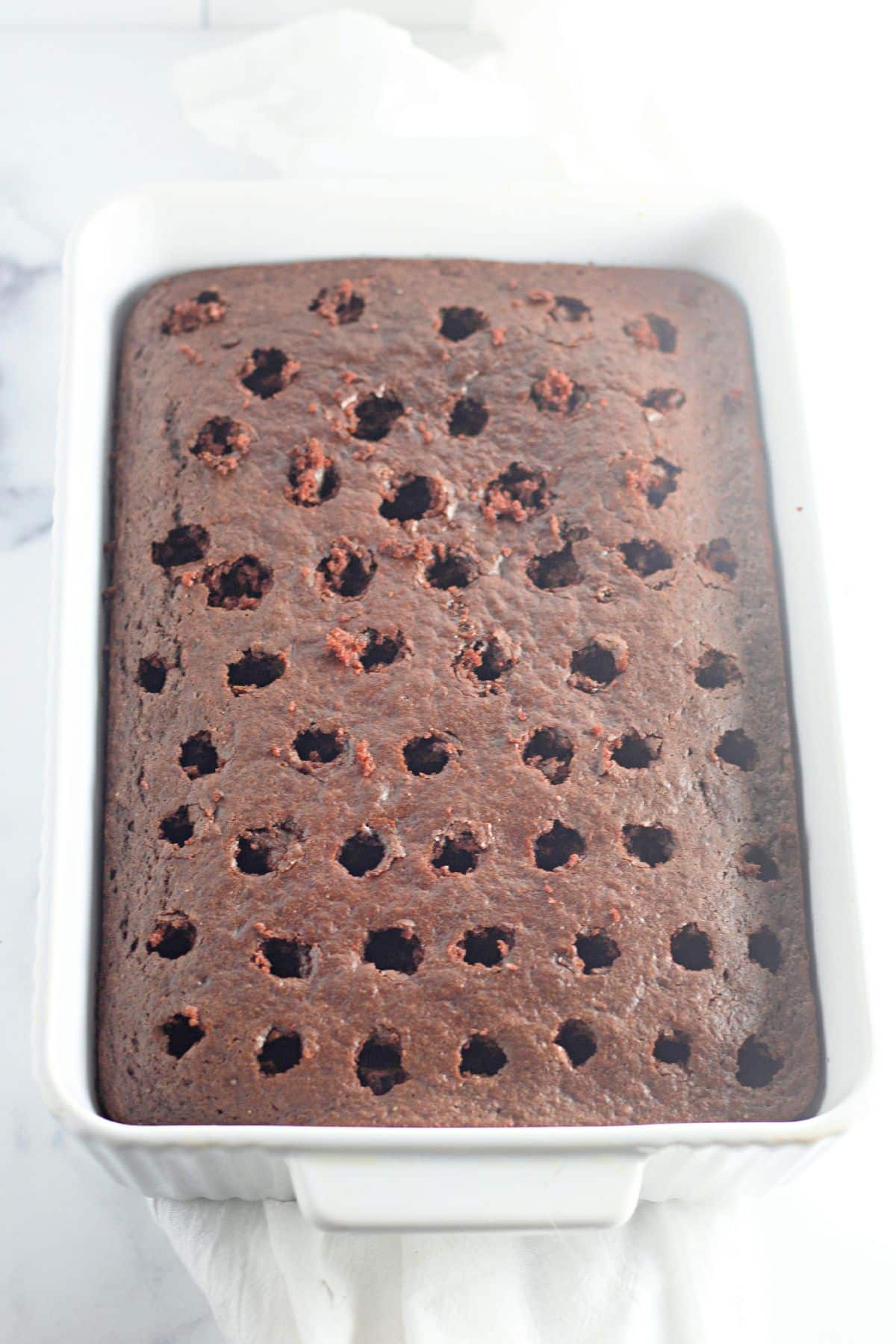 A chocolate sheet cake with evenly spaced holes poked in the top, in a white rectangular baking dish.