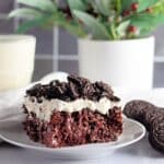 A slice of chocolate cake with cream and crushed cookies on a plate, with a glass of milk and plants behind.
