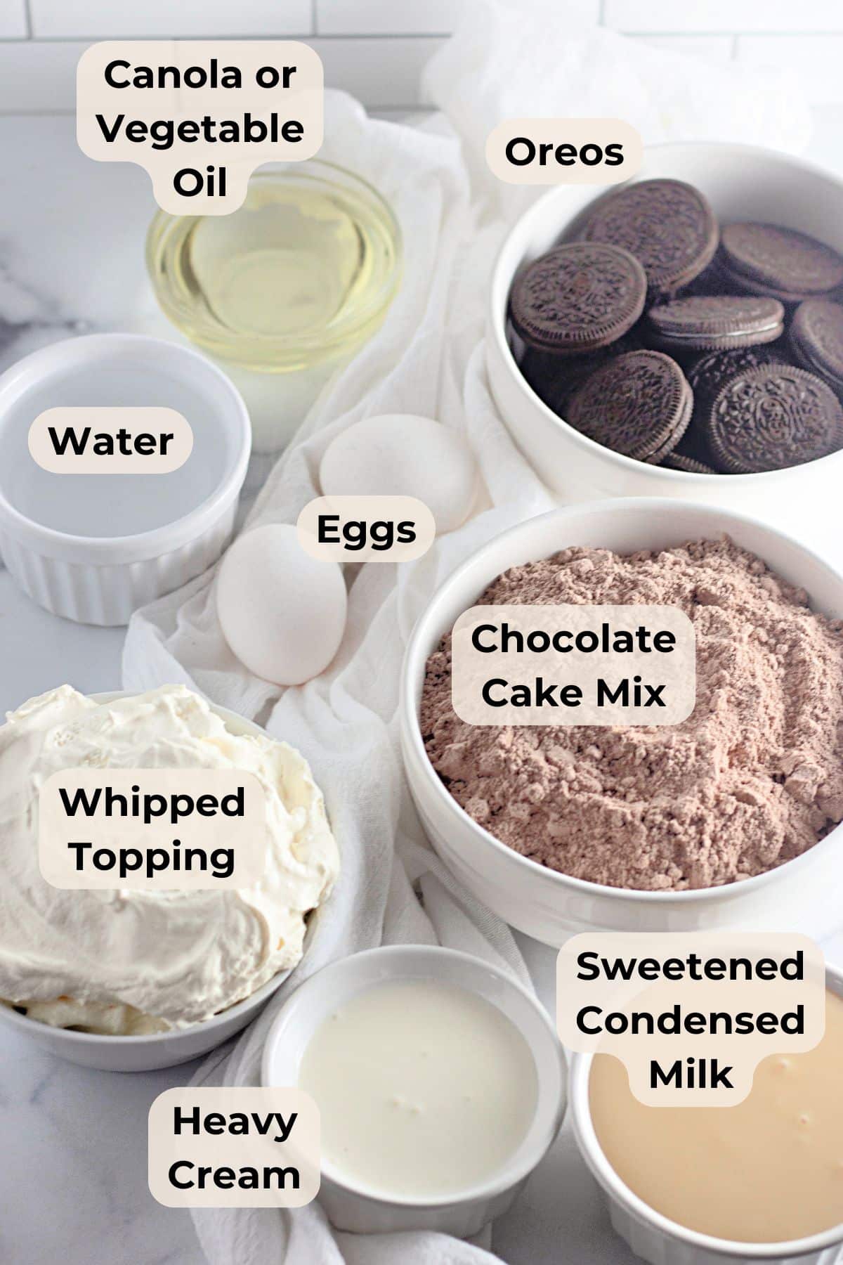 Bowls of canola oil, water, eggs, whipped topping, heavy cream, Oreos, cake mix, and condensed milk on a counter.