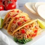 Three chicken tacos with shredded cheese and diced tomatoes, served with lime wedges on a white plate.