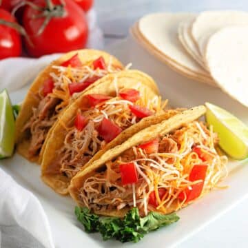 Three chicken tacos with shredded cheese and diced tomatoes, served with lime wedges on a white plate.