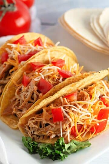 Three chicken tacos with shredded cheese and diced tomatoes, served with lime wedges on a white plate.