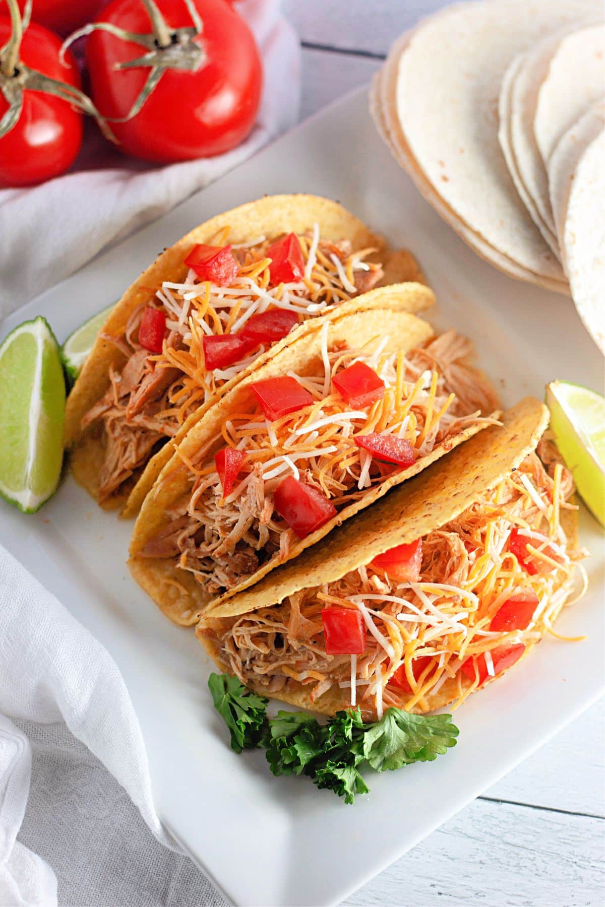 Three hard shell tacos filled with shredded meat, cheese, and diced tomatoes, served with lime wedges.