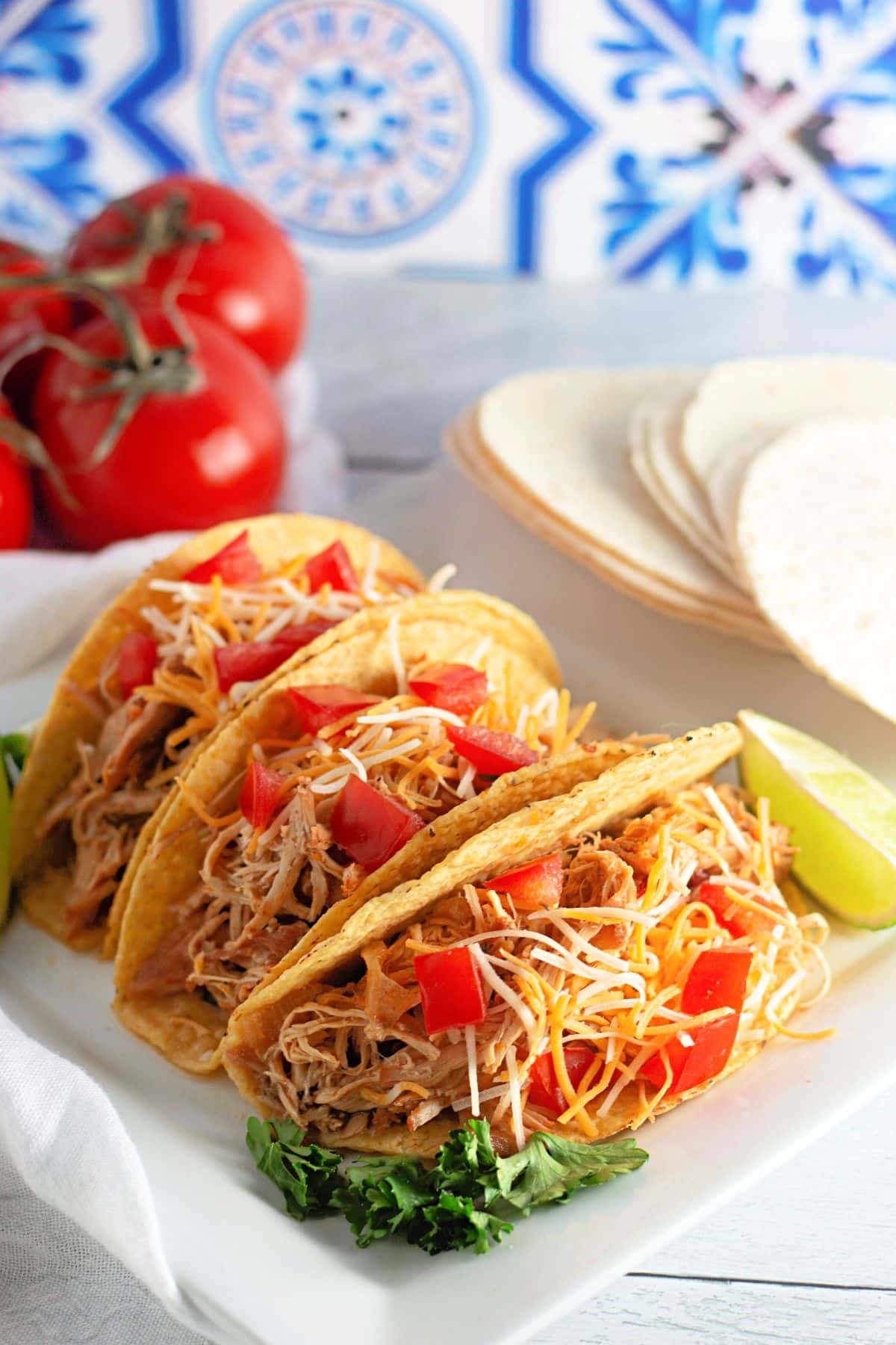Three shredded chicken tacos with cheese and tomato, served with lime, cilantro, tortillas, and fresh tomatoes.