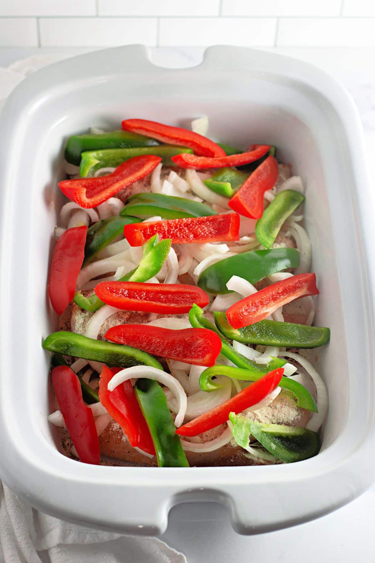 Sliced red and green bell peppers and onions in a white slow cooker, ready to be cooked.