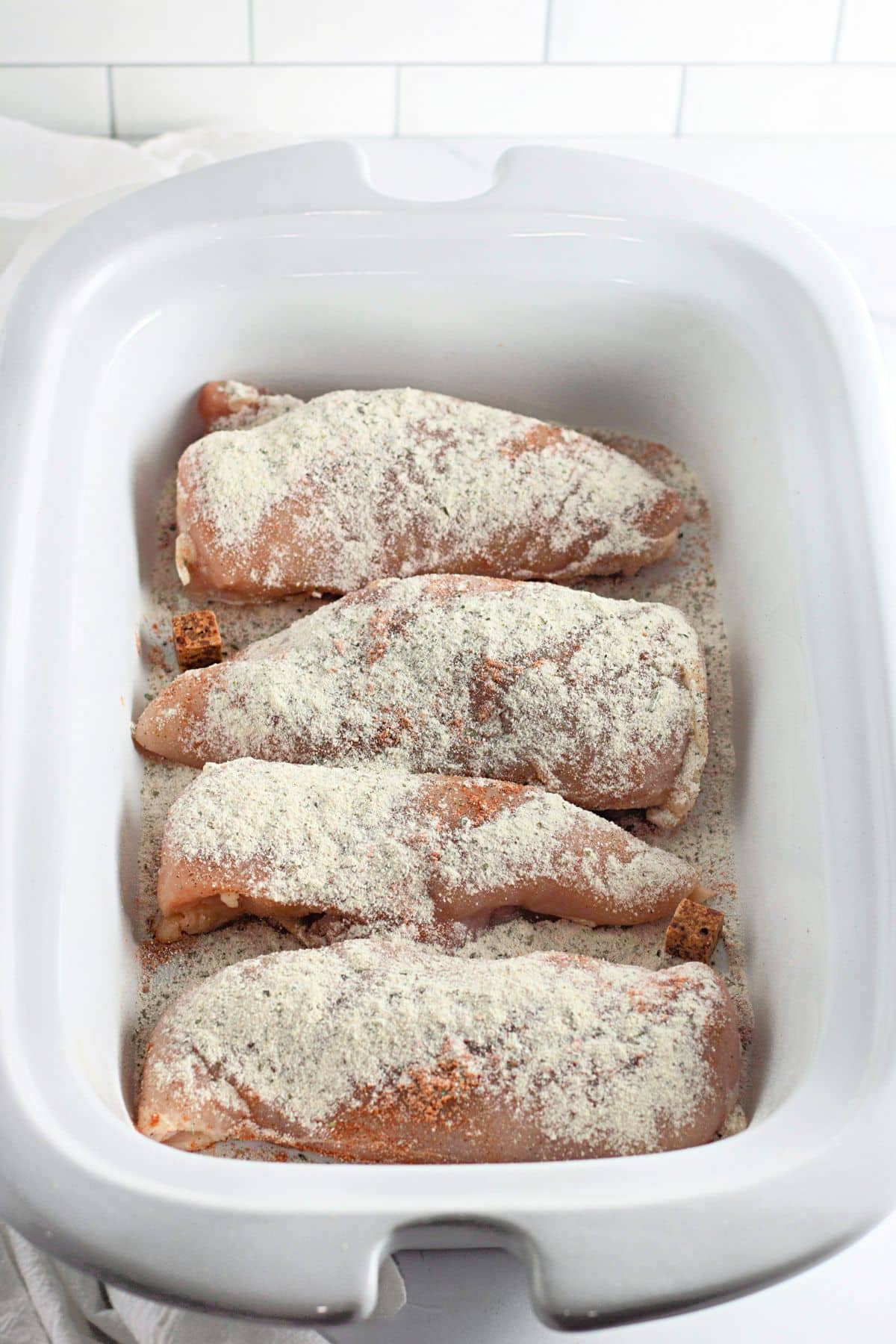 Four raw chicken breasts coated with a dry seasoning mix in a white ceramic slow cooker.