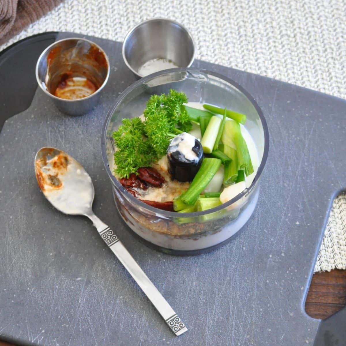 A food processor with herbs, green onions, and sauce sits on a dark cutting board with a spoon and dishes.