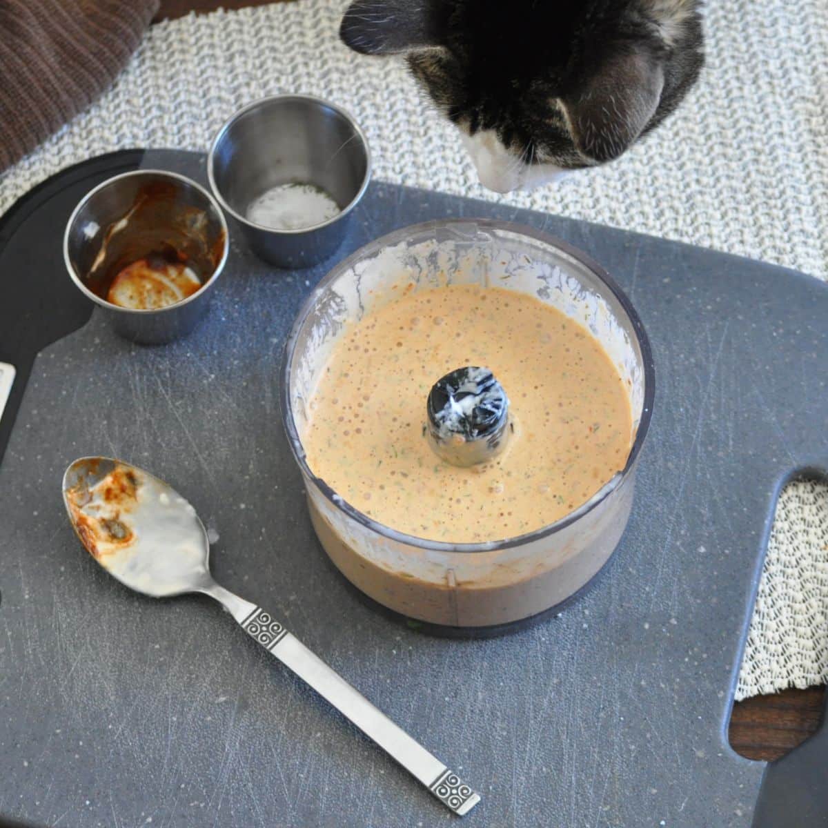 A cat looks at a food processor with sauce on a cutting board, next to a spoon and small bowls.