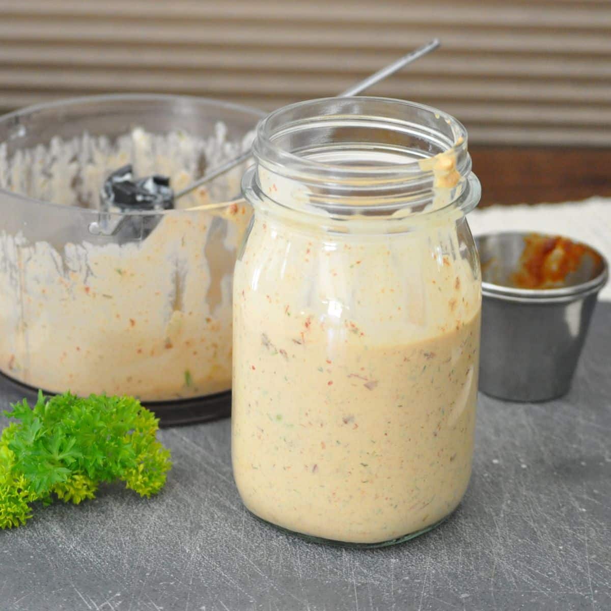 A jar of creamy, seasoned sauce sits on a counter near parsley and a food processor.